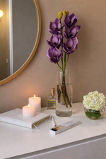 Vase with purple flowers on a table next to a candle and book, with a mirror and beige wall in the background.