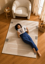 A woman in a navy blue jumpsuit lies on a cream and beige striped area rug on a hardwood floor, reading a magazine. The room features a white armchair, a small wooden side table, and a window with sheer curtains.