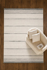 A rectangular cream and gray striped area rug laid on a dark wood floor, styled with a small cream upholstered accent chair and two matching pillows.