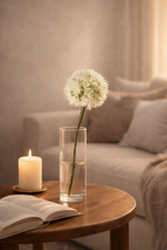 Flower in a vase on a wooden table with a candle and open book, against a neutral background.