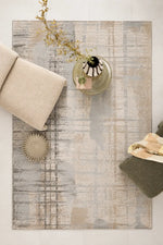 A rectangular area rug with a distressed, abstract pattern in beige, gray, and brown tones, styled in a modern living room setting with a beige sofa cushion, a glass vase with dried branches, and a dark green ottoman.