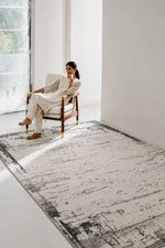 A woman in a white outfit sits in a wooden armchair beside a large rectangular area rug with a distressed black, gray, and off-white abstract pattern on a bright white room.