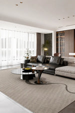A modern living room featuring a large light beige area rug under a black leather sectional sofa and round coffee tables, with large windows covered in white sheer curtains.
