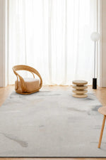 A large light gray and white abstract patterned area rug in a bright, modern room with a wicker chair, a small wooden side table, and a floor lamp.