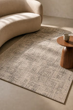 A rectangular area rug with a distressed abstract pattern in beige and gray, styled in a modern living room setting with a curved beige sofa and a small wooden side table.