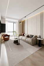 A modern living room featuring a light beige fabric sofa with dark accent pillows, a matching light beige area rug, a brown leather armchair, and a large window with a view of green trees.