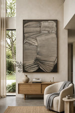A large framed abstract canvas print in grayscale tones hangs on a textured beige wall above a light wood console table in a modern, sunlit living room. The room features a cream-colored armchair with a gray throw blanket, a small side table, and a vase with dried branches.