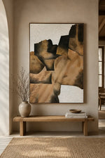 A framed abstract wall art print with geometric shapes in tan, brown, black, and off-white, displayed above a wooden bench in a warm neutral interior setting.
