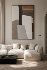 A large vertical abstract wall art print with geometric shapes in beige, brown, white, and black hangs above a cream-colored sectional sofa in a modern, sunlit living room.