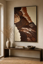 A large framed abstract wall art piece with layered brown, tan, and cream textures hangs above a dark wood console table in a styled interior with a vase, books, and a bowl.