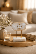 A small oval tabletop clock with a white marbled face and gold-tone frame, displayed on a marble-and-wood stand on a round wooden tray in a softly lit living room setting.
