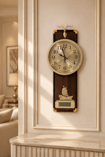 A vertical wall clock with a dark wood and gold-tone frame, featuring a cream-colored dial, a deer head topper, and a gold ship ornament, mounted on a cream-colored paneled wall in a warm, sunlit interior.