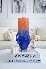 Blue and orange vase on books with a white couch and framed pictures in the background