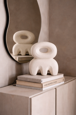 White abstract sculpture on books with a mirror reflecting another similar sculpture against a beige wall.