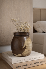 Brown ceramic vase with dried flowers on a stack of books in a room setting.