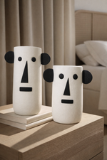 Two white vases with black facial features on a wooden surface.