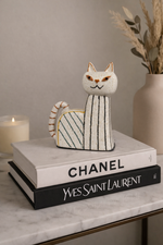 Cat-shaped decorative item on Chanel and Yves Saint Laurent books with a candle and vase in the background.