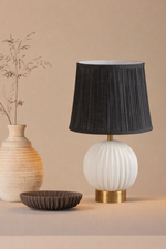 Table lamp with black pleated shade, white base, and gold accent on a beige surface.