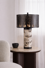 Table lamp with black textured shade on a round wooden table against a white wall.