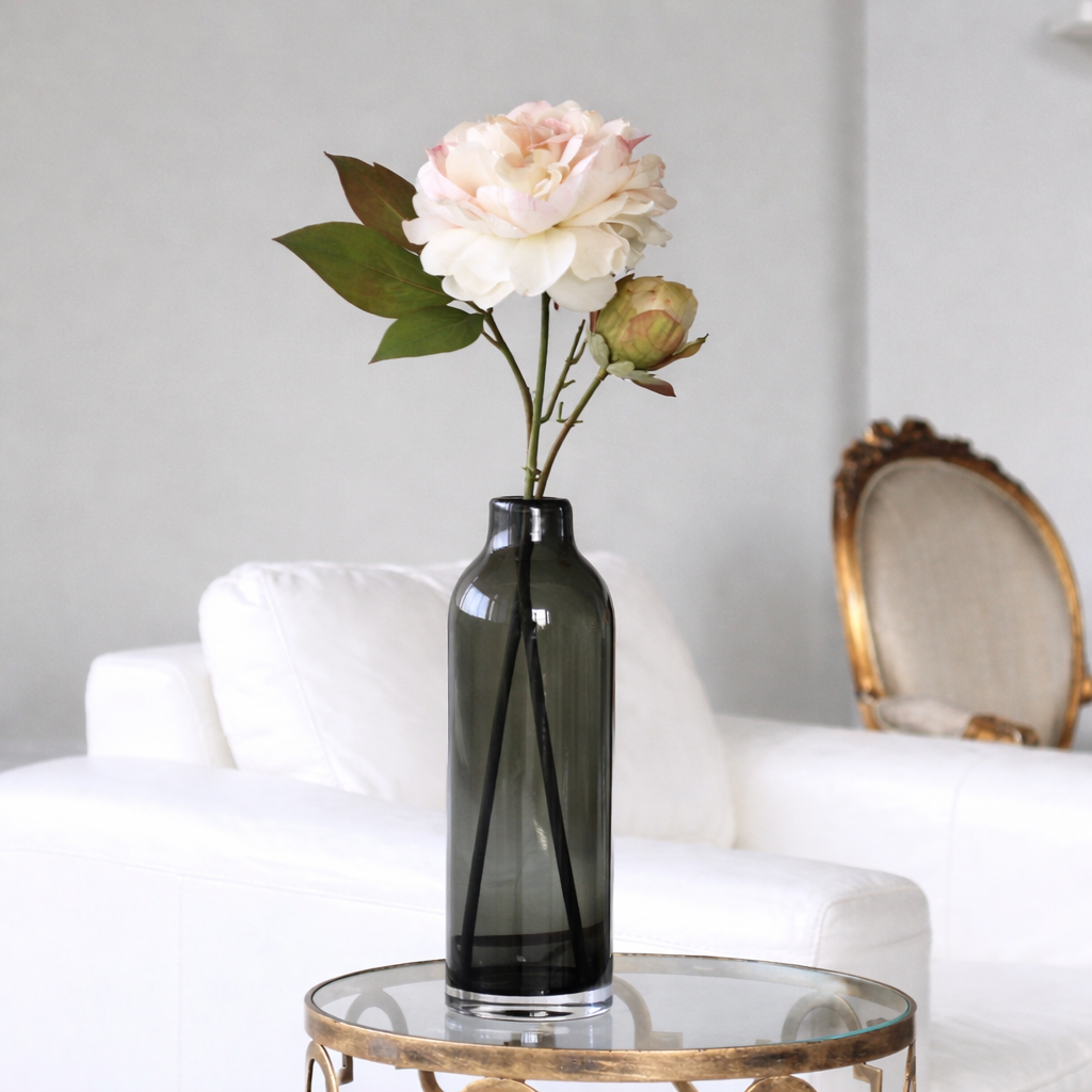 Dark glass vase with flowers on a round glass table in a room with a white armchair and decorative chair.