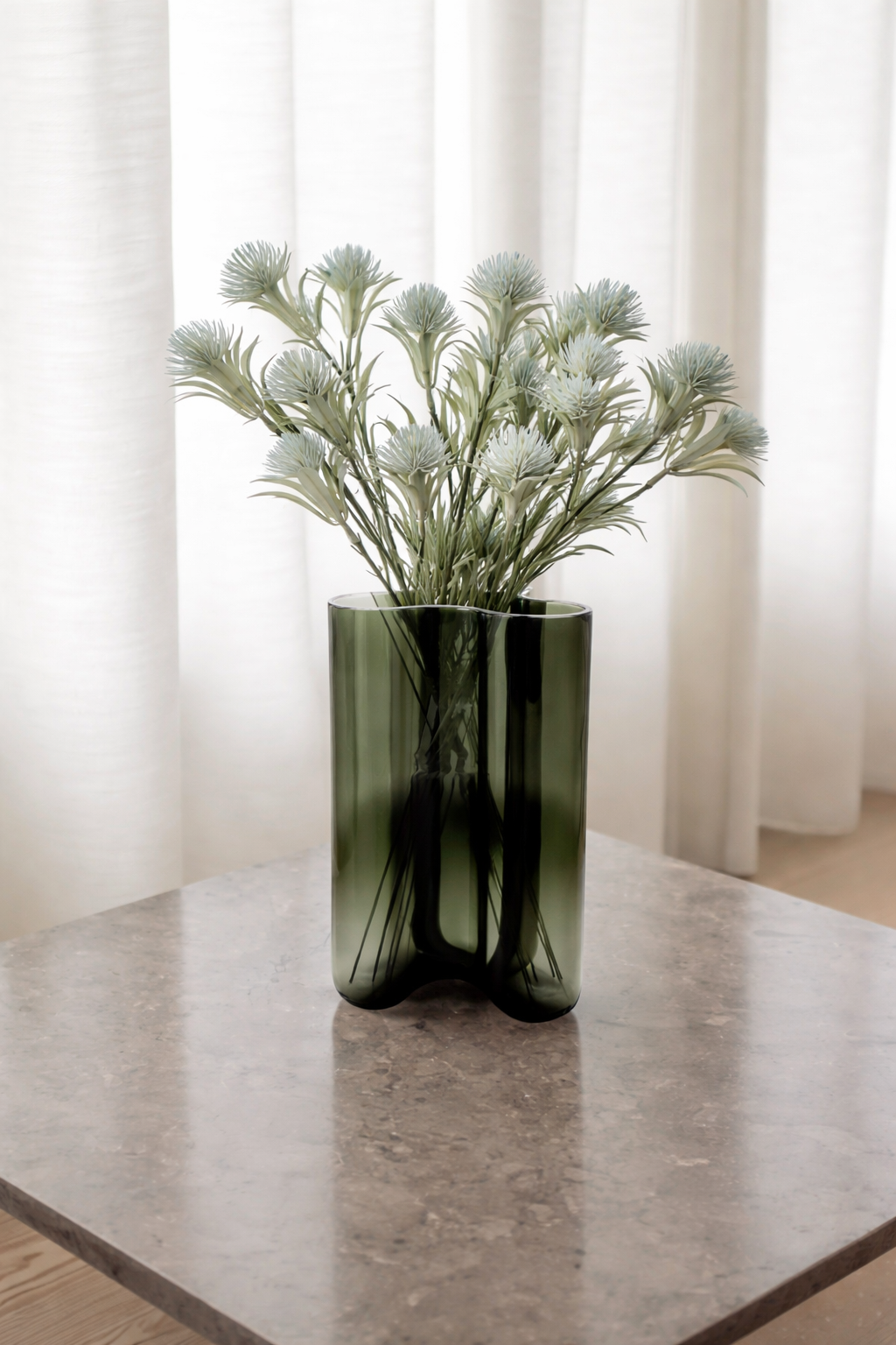 Green vase with white flowers on a marble surface with white curtains in the background