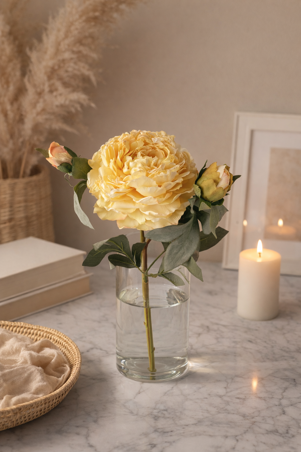 Yellow flower arrangement in a glass vase on a marble surface with a candle and books in the background.