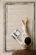 A top-down view of a woman in a light beige lounge set reading a magazine on a large beige area rug with a dark gray abstract border, in a cozy living room setting.