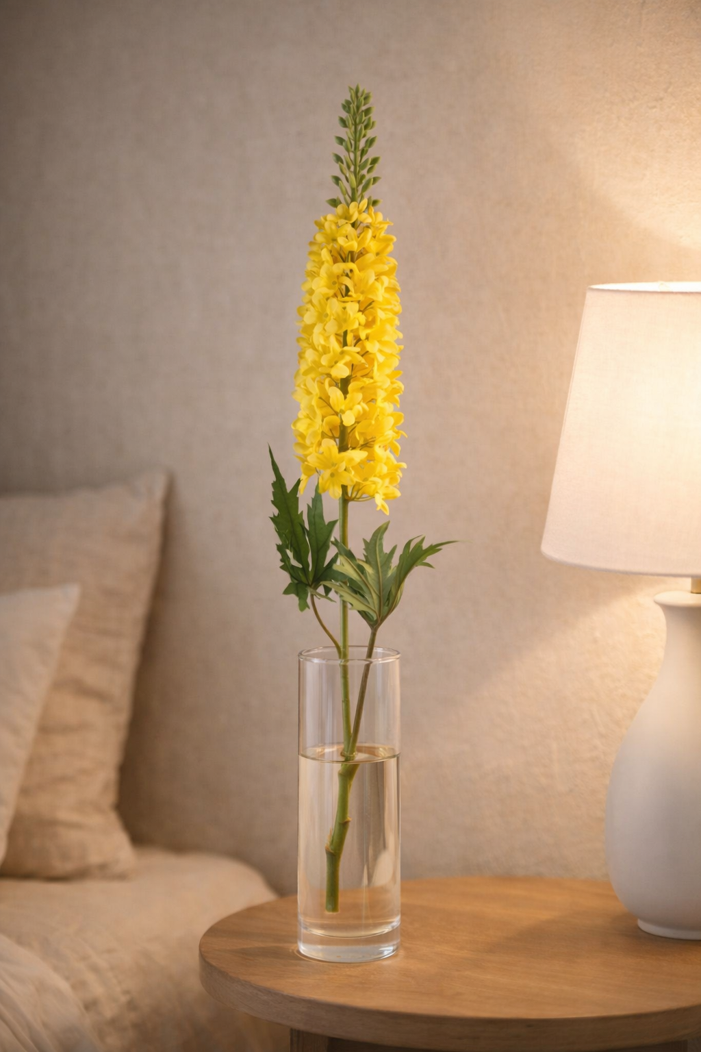 Yellow flower in a glass vase on a wooden table with a lamp and pillows in the background.