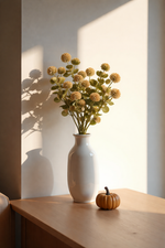 White vase with dried flowers on a wooden surface with a pumpkin, against a light wall.