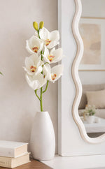 White vase with orchids on a wooden surface next to a mirror and books.