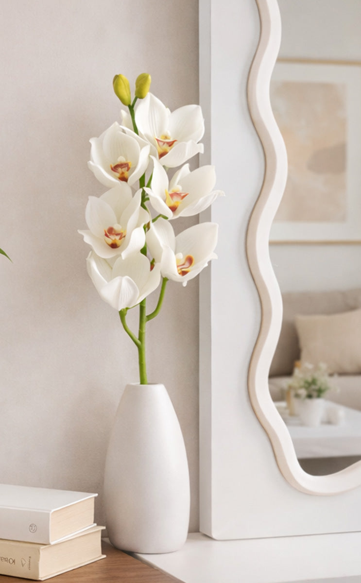 White vase with orchids on a wooden surface next to a mirror and books.