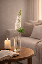 Vase with white flowers on a wooden table next to an open book and lit candle, with a sofa in the background.