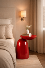 Red side table next to a bed with beige bedding and pillows, featuring a lamp and small plant.