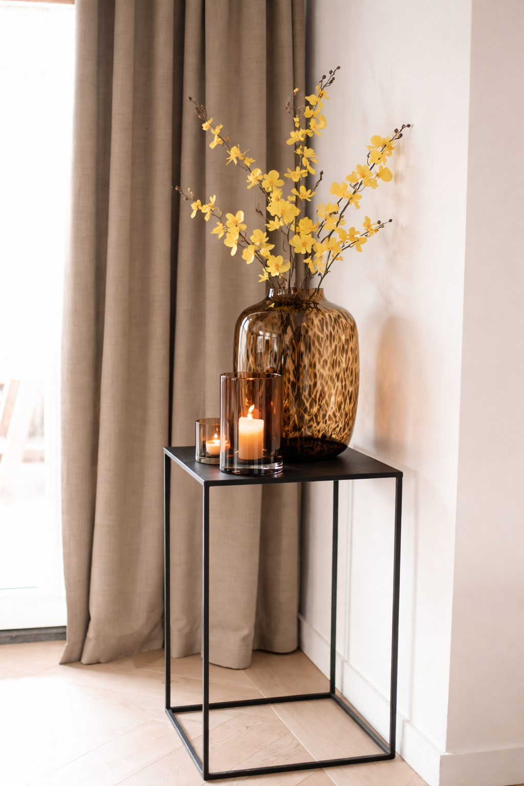 Decorative vase with yellow flowers on a small black table next to a window with beige curtains.