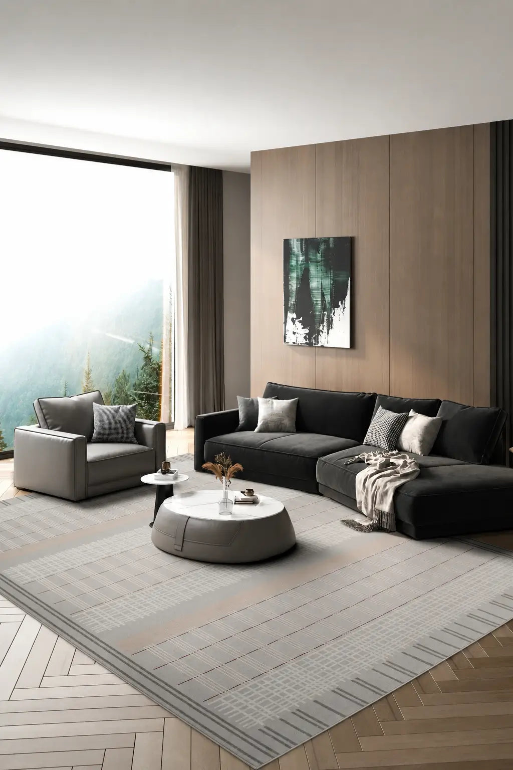 A modern living room featuring a dark gray sectional sofa, a matching armchair, a round gray ottoman, and a large textured light gray area rug on a wood herringbone floor. The room has a wood-paneled accent wall with a vertical window looking out to a misty mountain view.