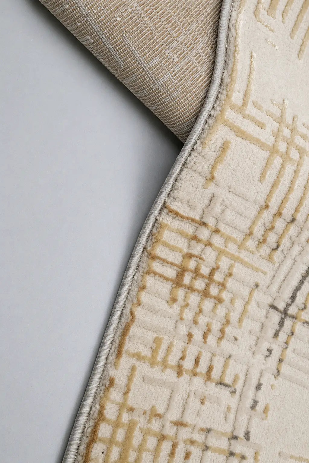 Close-up of a patterned area rug with a rolled corner showing the underside and edge, featuring a cream and beige geometric design on a light gray background.