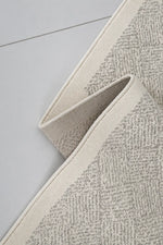 A close-up of a textured area rug with a herringbone pattern in beige and gray, folded over to show its woven texture and cream-colored edge.