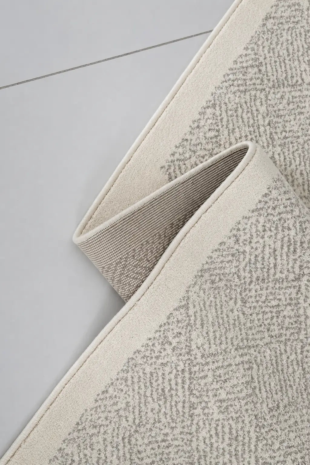 A close-up of a textured area rug with a herringbone pattern in beige and gray, folded over to show its woven texture and cream-colored edge.