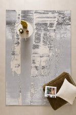 An abstract area rug with vertical brushstroke patterns in gray, white, and beige, styled in a modern living room setting with a side table, decorative objects, and a brown ottoman.
