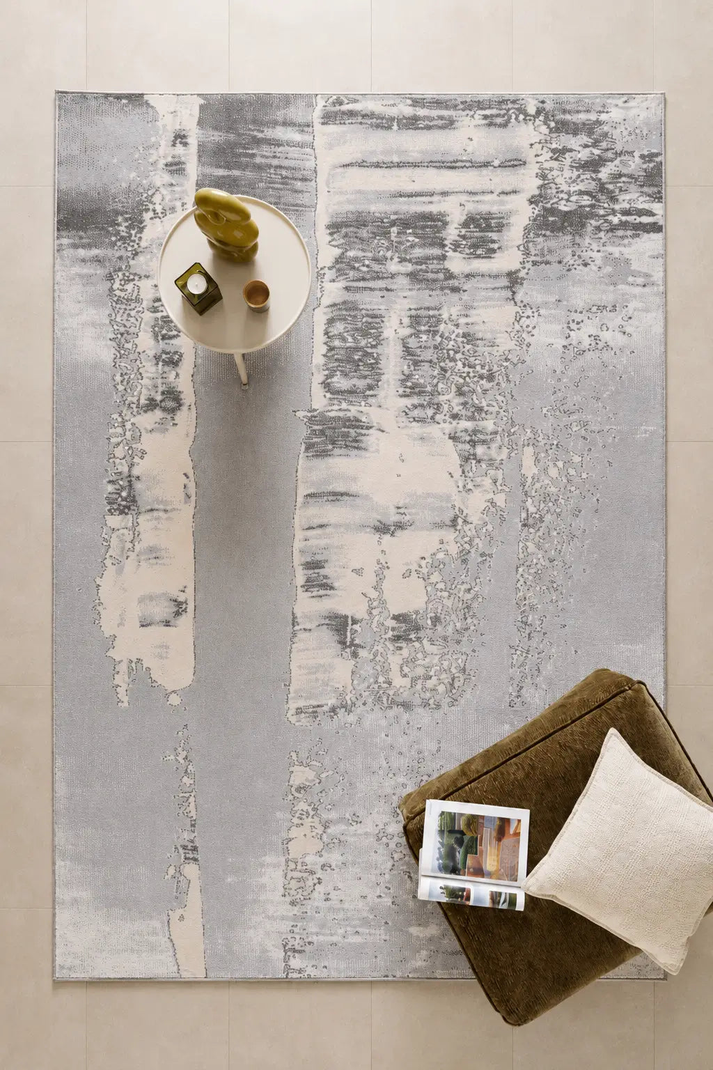 An abstract area rug with vertical brushstroke patterns in gray, white, and beige, styled in a modern living room setting with a side table, decorative objects, and a brown ottoman.