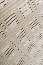 A close-up of a textured area rug featuring a modern abstract pattern of diagonal lines and dotted accents in dark brown on a warm beige and cream background.