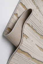 A close-up of a rolled area rug showing its textured beige and cream abstract pattern with subtle gray accents, set against a clean white background.