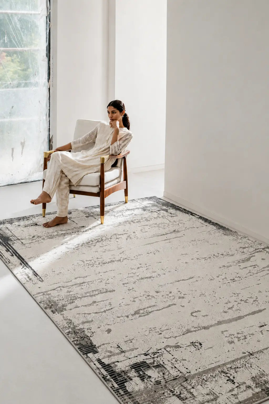 A woman in a white outfit sits in a wooden armchair beside a large rectangular area rug with a distressed black, gray, and off-white abstract pattern on a bright white room.