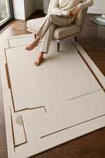 A large cream-colored area rug with a minimalist brown geometric line pattern, shown on a dark wood floor in a modern living room setting with a person sitting on a beige armchair.