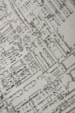 A close-up of a textured area rug featuring an abstract, distressed city-block pattern in shades of gray and black on a warm off-white background.