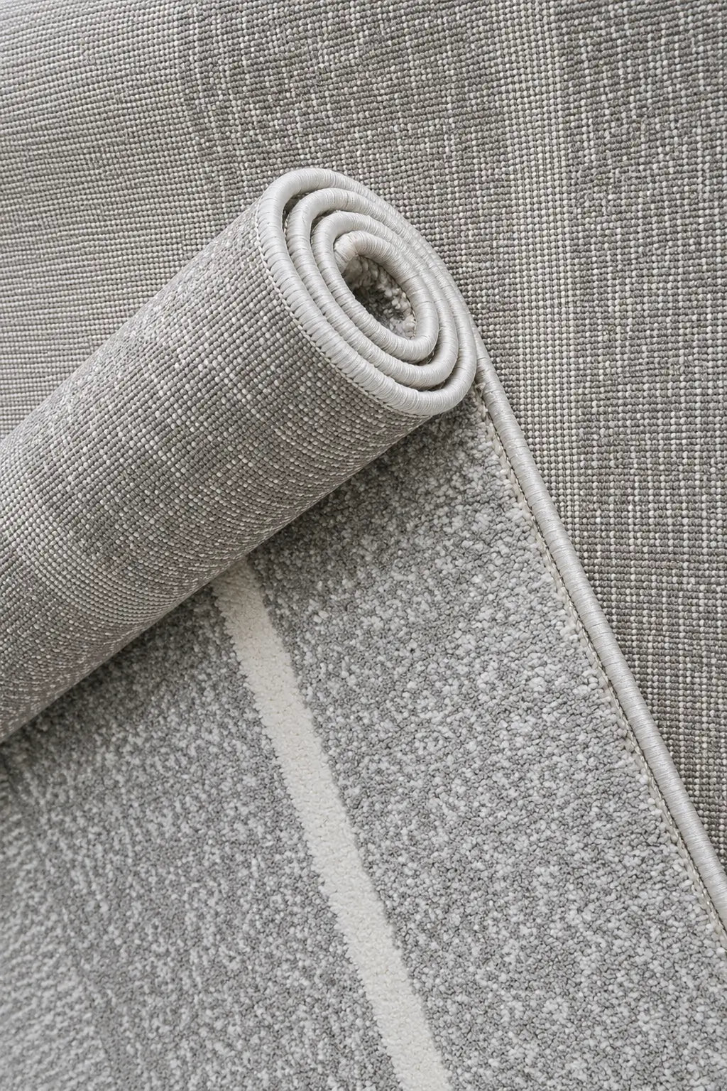 A close-up of a light gray textured area rug, partially rolled to show its pile and woven texture, with a subtle off-white stripe running along one side.
