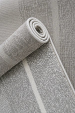 A close-up of a light gray textured area rug, partially rolled to show its pile and woven texture, with a subtle off-white stripe running along one side.