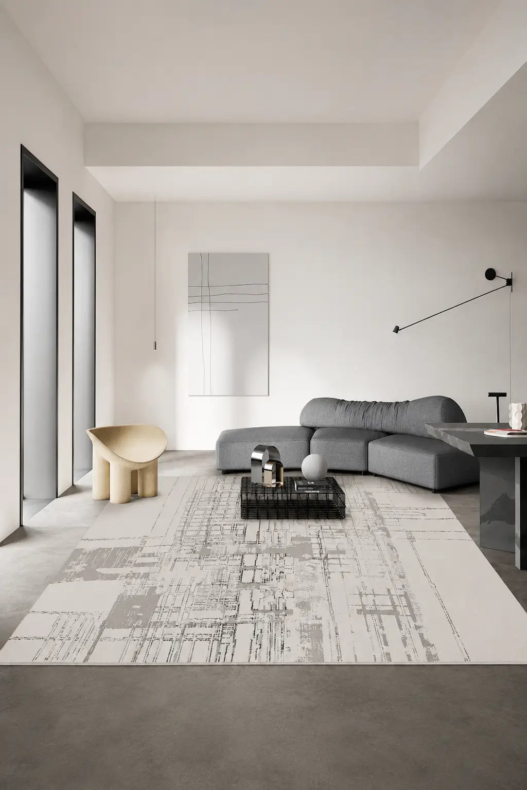 A modern living room featuring a large rectangular area rug with a gray and white abstract brushstroke pattern. The rug is styled in a minimalist room with a gray sectional sofa, a sculptural beige chair, and a black coffee table.