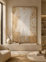 A large vertical abstract wall art piece with flowing white and gold brushstrokes, displayed in a warm, modern living room setting with a beige armchair, a round marble coffee table, and neutral-toned ceramic vases.