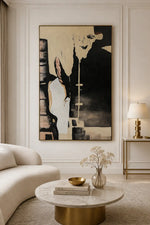 A large abstract canvas painting with black, beige, and white tones hangs on a white paneled wall in a modern living room setting with a curved cream sofa, a marble-top coffee table, and a gold side table with a lamp.